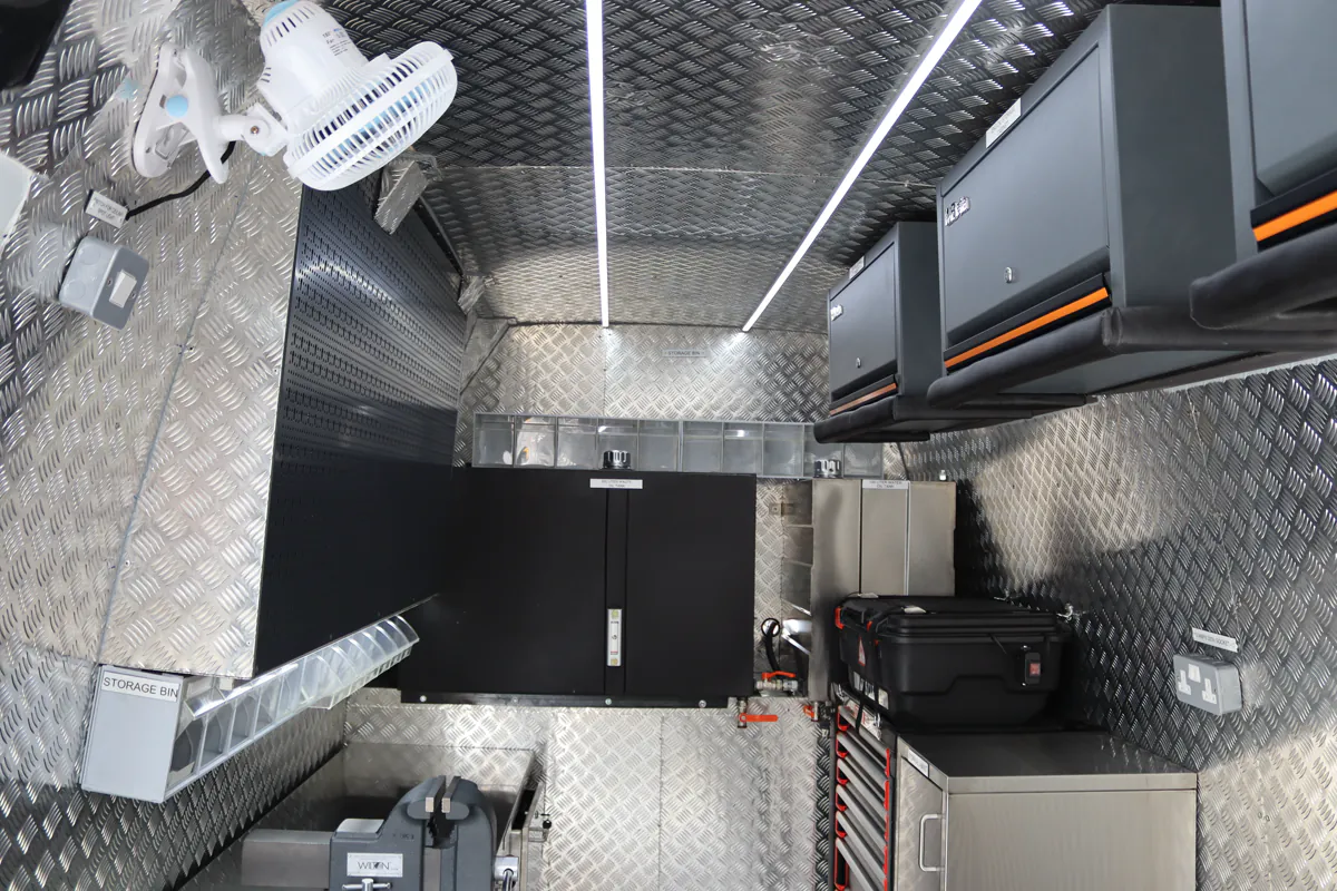 Interior of a fitted workshop cabin with chequer-plate lining, overhead cabinets, ceiling lighting and floor-mounted equipment